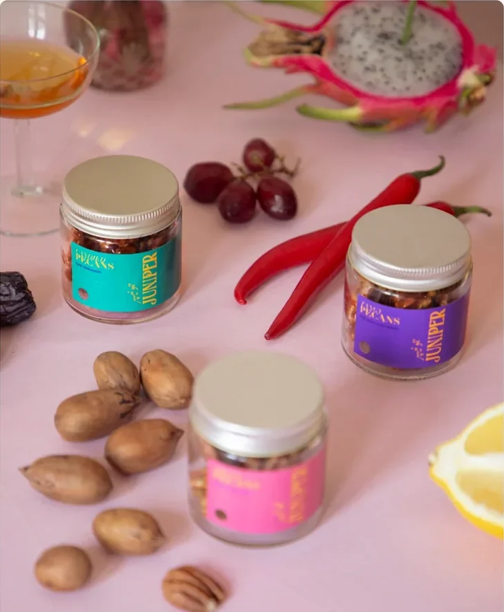 Three glass jars of pecans with teal, purple and pink labels on a pink table, among whole pecans, red chillies, grapes, a lemon and half a dragon fruit