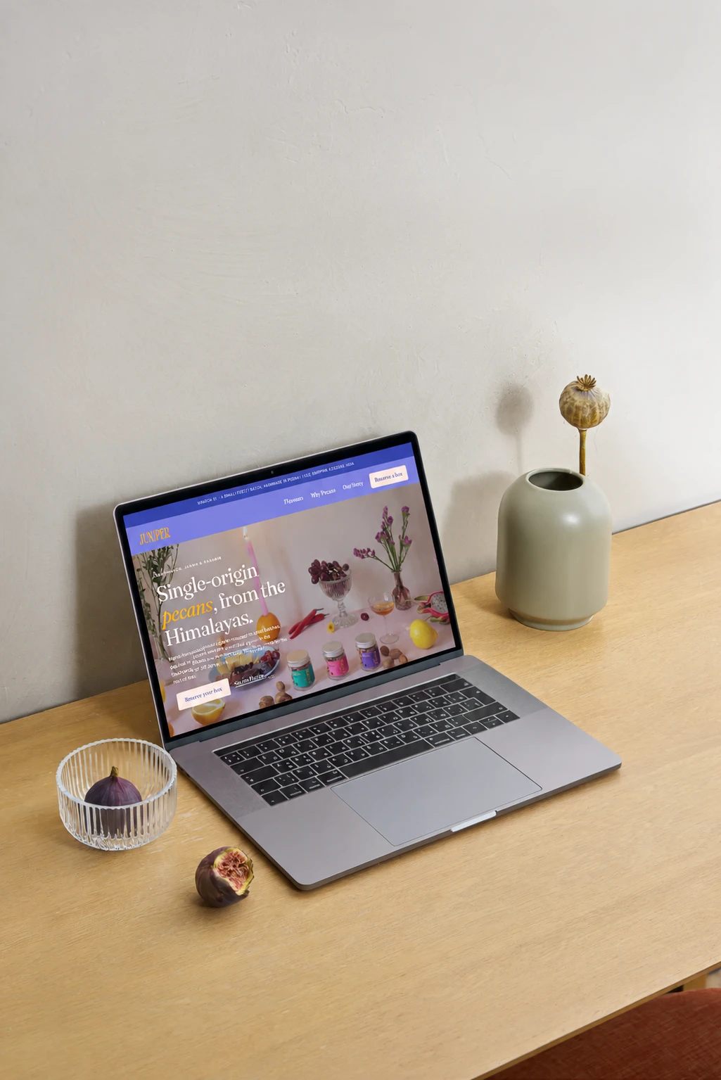 A laptop angled on a light wooden desk against a plaster wall, showing the Juniper home page, with a glass bowl holding a fig and a dried poppy head in a sage vase beside it